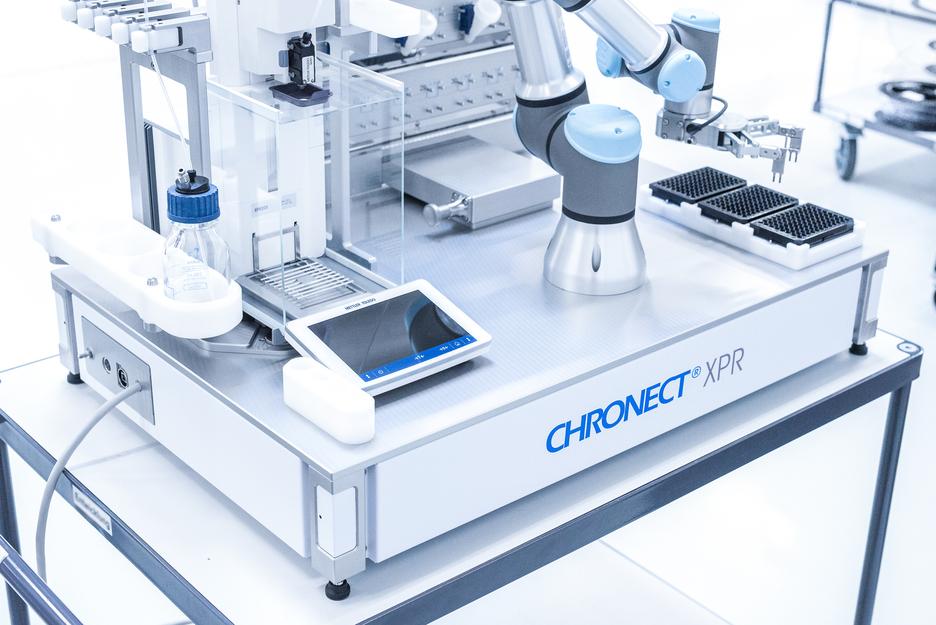 Automatisiertes Laborsystem mit Roboterarm, Tablet und Behälter auf einem Tisch in einem modernen Labor.