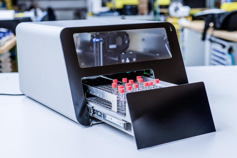 Moderne Labormaschine mit ge&ouml;ffnetem Fach f&uuml;r Glasfl&auml;schchen auf einem Tisch in einem technischen Umfeld.