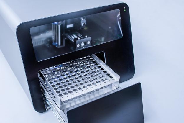 Moderne Laborgeräte mit einer herausziehbaren Metallablage für Proben im Vordergrund, hinter Glas sichtbare technische Komponenten.